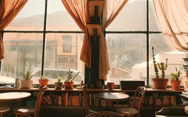 Mese rotunde din lemn lângă o fereastră mare, cu perdele și cactuși pe pervaz