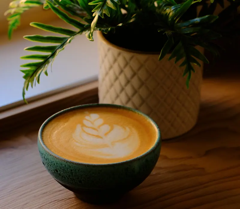Latte cu rozetă într-un bol verde, pe o masă de lemn, lângă o plantă